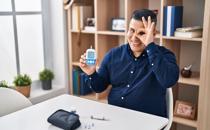 A man holding a blood glucose meter, looking through a hand gesture framing his eye